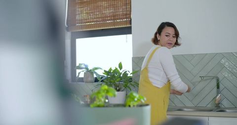 Asian woman washing dishes in bright modern kitchen with plants and yellow apron