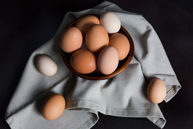 Farm fresh brown and white eggs are placed in a wooden bowl, with a gray fabric underneath and scattered around. This rustic setting captures the essence of organic and natural food. Great for promoting clean eating, nutrition articles, and culinary blogs.