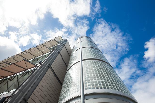 This image showcases a modern skyscraper with a glass facade against a bright blue sky with scattered clouds. Ideal for use in business presentations, real estate promotions, architectural portfolios, and urban development projects. The low angle perspective emphasizes the height and grandeur of the building, making it suitable for themes related to growth, progress, and modernity.