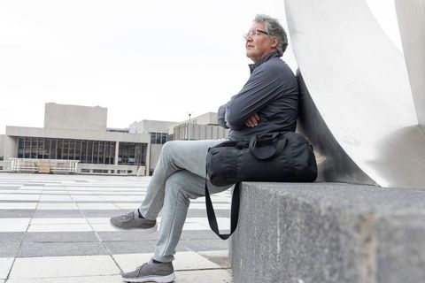 Contemplative senior man sitting on urban plaza with duffel bag and modern sculpture