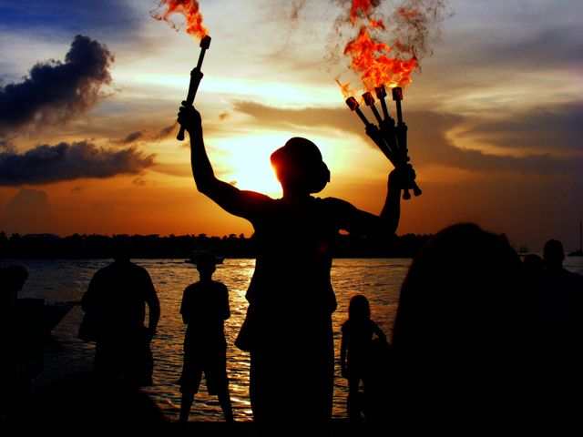 Silhouette of a fire performer holding up flaming torches against a vibrant sunset over the ocean, with other silhouettes visible in the background. Ideal for use in promotions for beach events, travel brochures, performance arts advertisements, cultural events, or any content associated with entertainment and dramatic scenes.