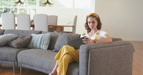 Young woman wearing yellow overalls sitting on gray sofa using tablet in bright open-plan room