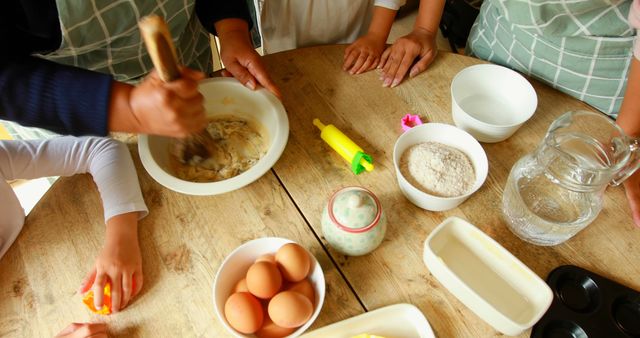 Family engaging in baking activity around wooden table, hands visible mixing ingredients. Various baking items like eggs, flour, bowls emphasize family bonding and teamwork. Great for illustrating home cooking, lifestyle, family activities, or children's participation in cooking.