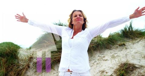 Mature woman stretching arms on coastal dunes wearing white linen with upward arrow overlay