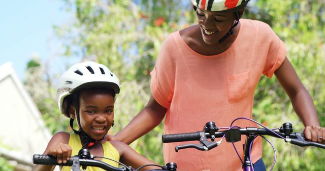 Content depicts a joyful mother helping her young daughter learn how to ride a bicycle in a sunny park. Both are wearing helmets, emphasizing safety during this outdoor activity. This can be used in articles or advertising for topics like parenting, outdoor activities, family bonding, and child safety.