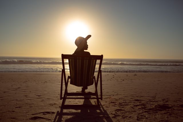This image depicts a woman sitting in a beach chair, gazing at the ocean during sunset. The silhouette effect adds a sense of tranquility and contemplation. Ideal for travel brochures, vacation advertisements, relaxation and wellness content, and inspirational blogs.