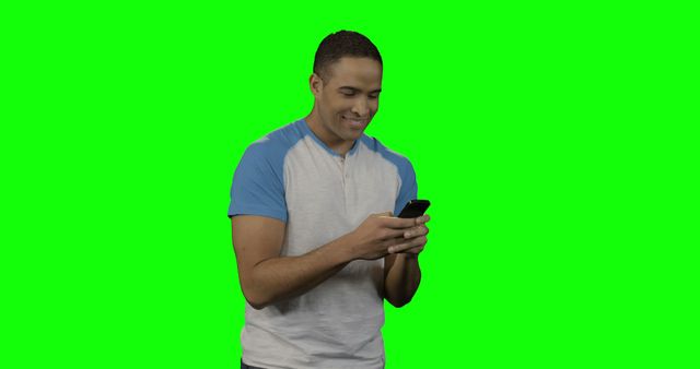Young man wearing casual clothing using smartphone against green screen background. He appears to be smiling and engaged. Ideal for graphics and visual effects projects. Suitable for technology, communication, and lifestyle promotions.