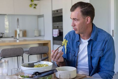 Middle-aged man enjoying pesto pasta at bright modern kitchen table, casual home dining
