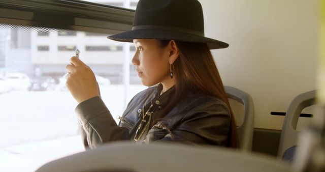 Fashionable asian woman wearing hat sitting on bus using smartphone on sunny day. Communication, street fashion, city living, transport and lifestyle, unaltered.