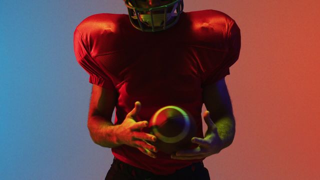 American football player stands under dramatic neon lighting, offering a dynamic and vivid portrayal of sport and competition. The essence of action is captured with an intense red-to-blue gradient providing a lively contrasting backdrop. Useful for sports-themed promotional materials, advertisements, or illustrating intensity and focus in athletics environments.