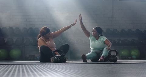 High-fiving diverse women after kettlebell workout sitting on gym mat in moody light