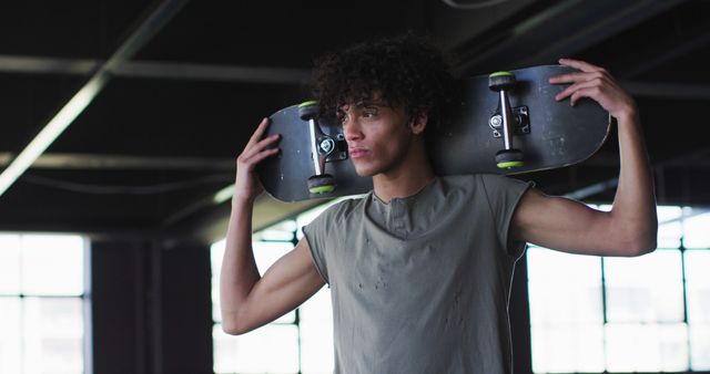 Young man holding skateboard on shoulders, standing in modern indoor skate park. The image conveys an urban lifestyle, casual attitude, and active youth culture. Ideal for use in advertising related to sports, skateboarding, streetwear, youth activities, and urban living.