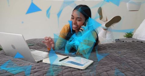 Young woman working remotely on bed talking on phone typing on laptop with notebook and pen