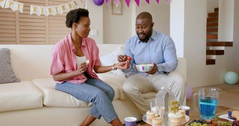 African American Couple Celebrating Cozy Home Birthday Sharing Cupcakes on Sofa with Balloons