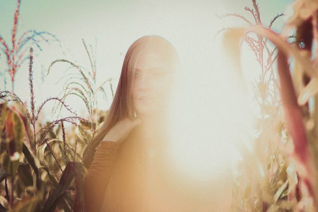Young woman is standing in cornfield during golden hour, light creating a dreamy glow. Perfect for themes of nature, relaxation, farmhouse lifestyle, or summer fashion. Great for magazines, blogs, or advertisements focusing on rural living, appreciating nature, or female empowerment.