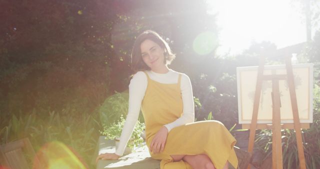 Young woman in yellow dress sitting in lush green garden with bright sunlight. Perfect for lifestyle blogs, fashion editorials emphasising summer styles, or promotion of outdoor activities. Creates relaxed and enjoyable mood with sunlight adding a dreamy, hazy effect.