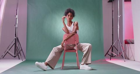 Confident female model posing on pink chair against green backdrop wearing casual fashion
