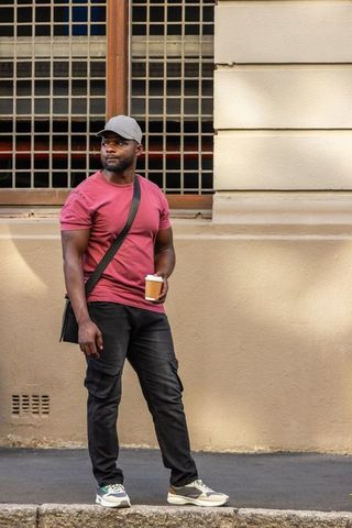 Urban casual commuter standing with coffee and satchel by gridded window in sunlit street