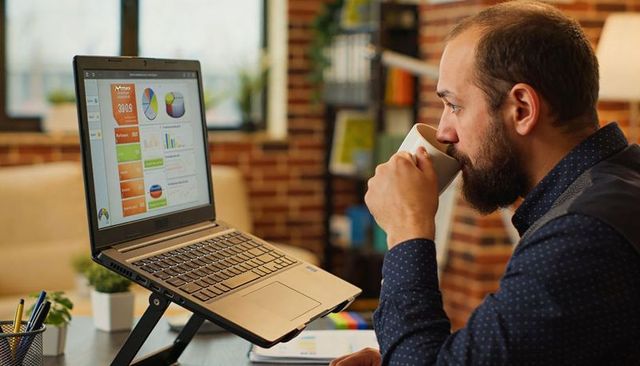 Bearded man sipping coffee while reviewing business analytics on laptop stand in loft