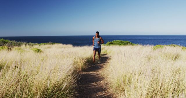 Man running on a coastal trail lined with tall grass under a clear, blue sky. Ocean in the background, sunlight creating a serene environment. Perfect for promoting outdoor fitness, active lifestyle, nature exploration, sports and wellness articles, or coastal tourism.