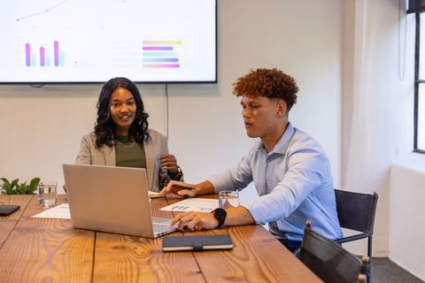 Diverse coworkers analyzing data on laptop and large screen in modern boardroom