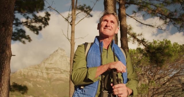 Elderly man enjoys hiking in mountainous forest area with sunlight filtering through trees. Man wears green shirt, blue vest and carries binoculars and backpack, indicating a love for nature, travel, and active lifestyle. Perfect for depicting adventure, healthy pastimes for seniors, outdoor activities, and exploration in marketing or informational content.