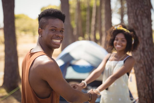 Young couple enjoying a camping trip in a forest, holding hands and smiling. Ideal for use in travel blogs, outdoor adventure promotions, relationship and bonding themes, and summer activity advertisements.
