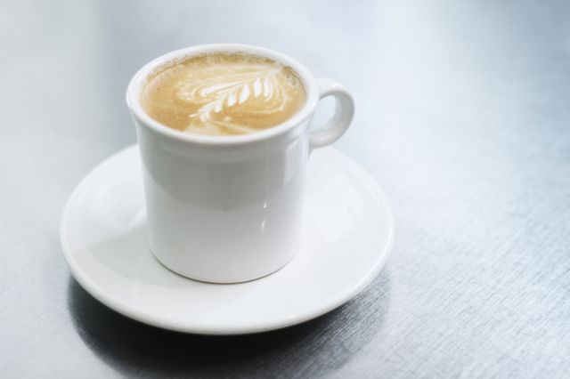 White coffee cup with foam art on saucer on metal surface. Perfect for use in cafe menus, coffee shop advertising, morning routines, blog posts, and social media content focused on coffee culture.