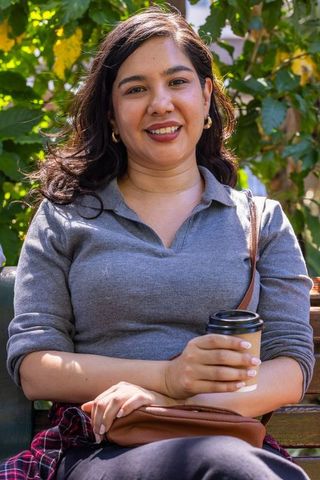 Smiling woman holding coffee on park bench, sunlit outdoor casual lifestyle portrait