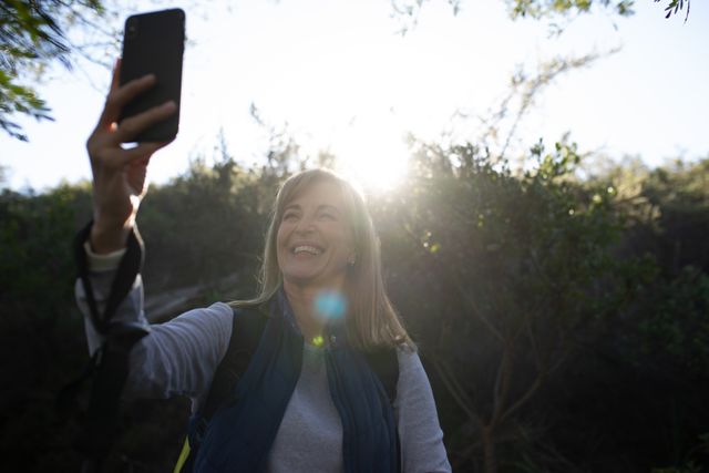 Front view of a happy senior Caucasian woman enjoying time in nature, hiking in mountains on a sunny day, smiling and taking a selfie with her smartphone