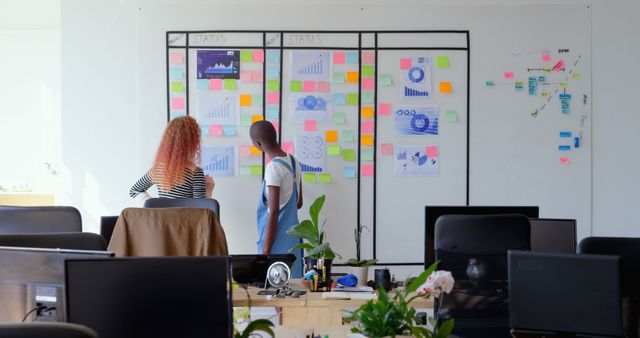 Coworkers at a planning board collaborating in a modern open office space. Sticky notes, graphs, and charts are displayed on the board indicating various project statuses. Perfect for depicting business planning, teamwork, modern office environments, and organizational development. Useful for website banners, blogs, articles on teamwork, project management, and office productivity concepts.