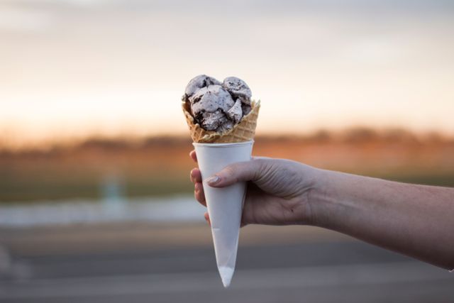 Hand holding ice cream cone outdoors with a sunset backdrop. Perfect for advertisements related to desserts, outdoor activities, summer events, and food promotions. Great for social media posts and food blogs.