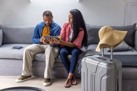 Couple planning trip and booking travel on tablet at home next to suitcase and sun hat