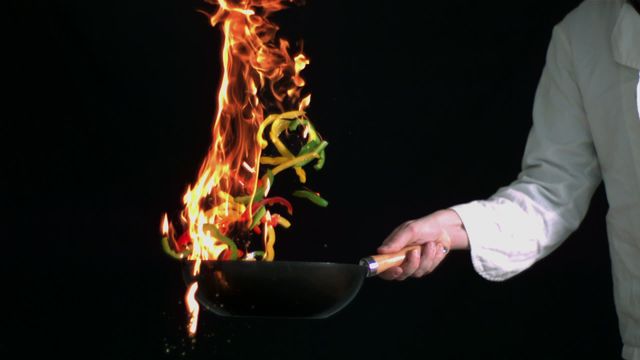 Chef is tossing and flambéing sliced peppers in a flaming pan against a black background, showcasing high-level cooking techniques. This visually striking and dramatic scene is perfect for illustrating articles or promotions about advanced culinary skills, cooking demonstrations, or creative cooking recipes. It emphasizes excitement and boldness in the culinary arts.