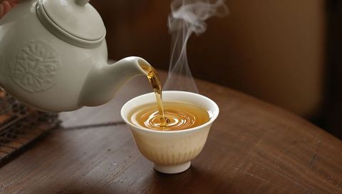 Pouring amber tea into porcelain cup with rising steam on wooden table