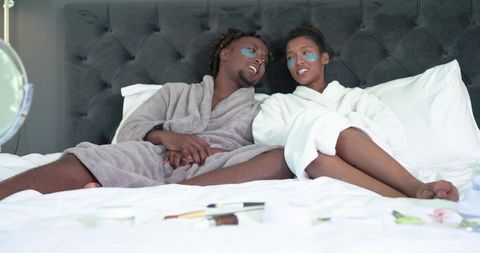 African american couple reclining in bed wearing robes and under-eye patches sharing relaxation
