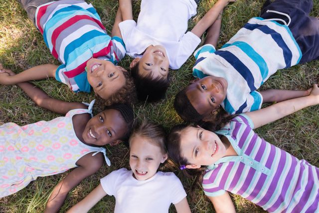 This image shows a diverse group of children lying on the grass in a circle, smiling and enjoying each other's company. Perfect for use in educational materials, advertisements promoting diversity and inclusion, summer camp brochures, and articles about childhood friendship and outdoor activities.