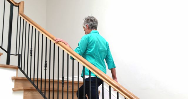 Back view of caucasian senior woman in blue blouse walking on stairs at home, copy space. Retirement, domestic life and senior lifestyle, unaltered.