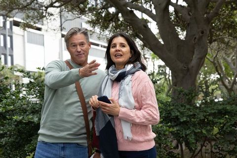 Middle-aged couple exploring urban park, pointing directions, holding smartphone, wearing sweaters