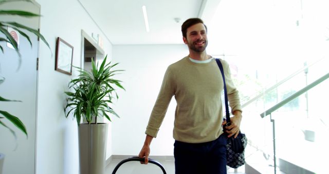 Man in comfortable attire smiling and holding luggage in modern office lobby filled with daylight and plants. Ideal for concepts related to business travel, corporate lifestyle, workplace, modern office environments.