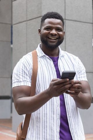 Smiling man texting on smartphone in urban plaza wearing striped shirt and shoulder bag