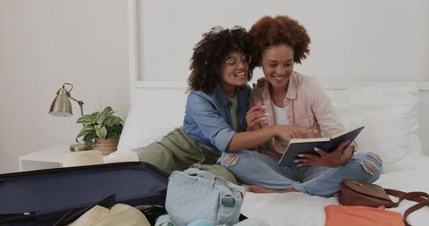 African American Women Packing and Planning Vacation While Reading Travel Journal on Bed