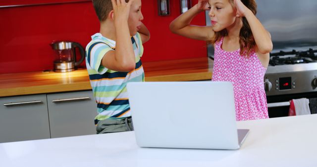 Two children, a boy and a girl, appear frustrated or confused while looking at a laptop screen in a kitchen setting. Their expressions suggest they might be dealing with a challenging task or unexpected problem on the computer.