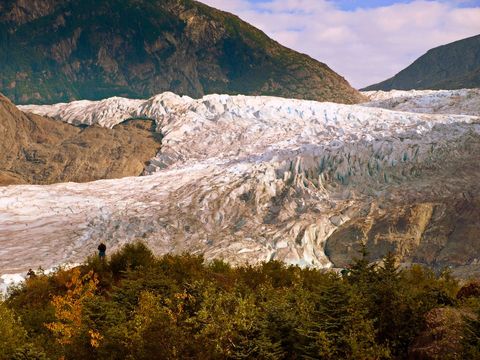 Hiker watching vast glacier flowing through rugged mountain valley with autumn foreground
