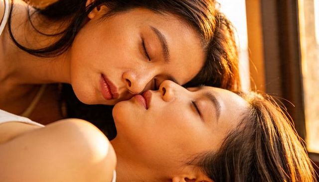 Two asian women sharing tender sunlit closeup, leaning faces at window, intimate couple cuddling