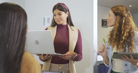 Female coworkers presenting laptop and taking tablet notes in modern office corridor