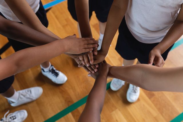 This image depicts a group of diverse schoolchildren forming a hand stack in a basketball court, symbolizing teamwork and unity. It is ideal for use in educational materials, sports promotions, teamwork and collaboration campaigns, and articles about childhood development and physical education.