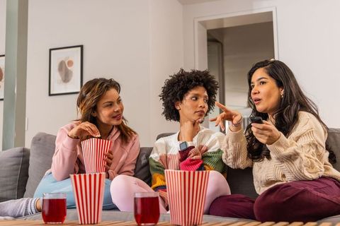 Diverse women sharing popcorn and watching tv on cozy sofa during at-home movie night