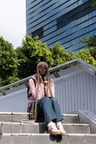 African american student sitting on city steps wearing pink beanie and headphones