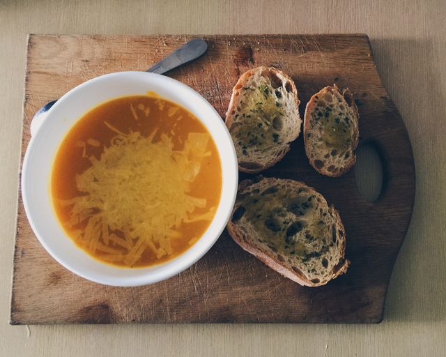 Tomato soup with grated cheese and slices of garlic bread on wooden board creating homely, comforting food presentation. Ideal for food blogs, recipe websites, or menu designs displaying savory, homemade meals.